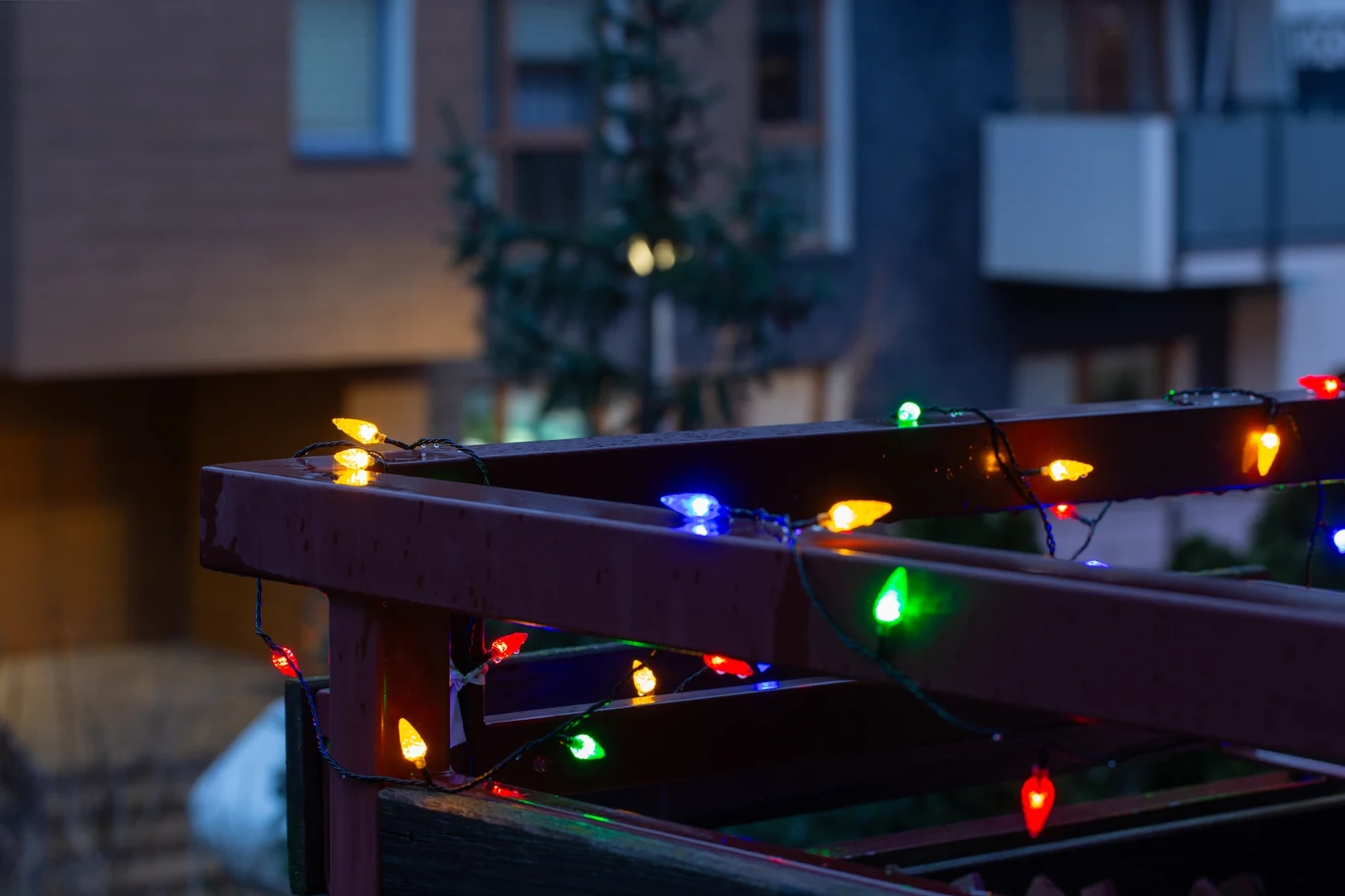 Spalvotos kalėdinės lemputės apšviečia medinę konstrukciją daugiabučių namų fone, kurdamos jaukią šventinę atmosferą.